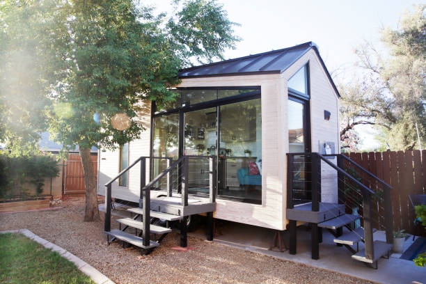 A tiny house with large glass windows, sits in the backyard surrounded by a wooden fence and trees.